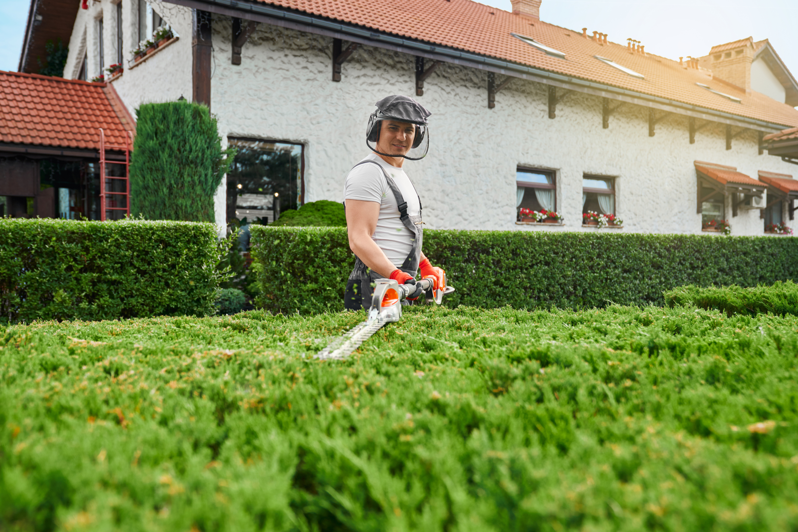 Handsome young man in working uniform, protective glasses and gloves pruning green bushes at garden during summer time. Concept of seasonal work and landscaping.